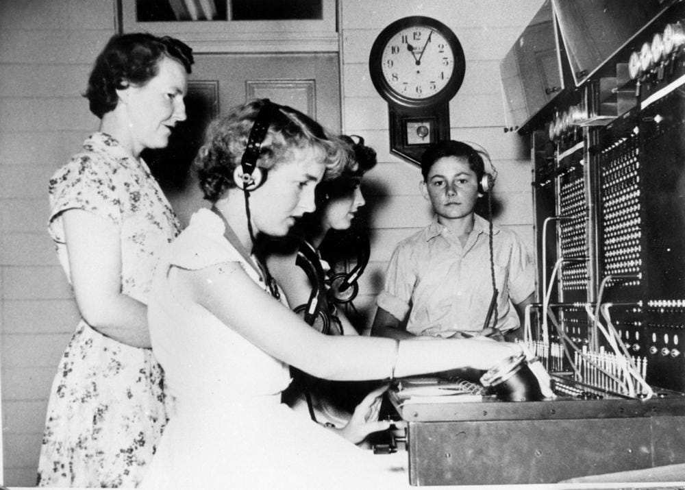 Deux téléphonistes, dont une casquée, travaillent devant un central téléphonique tandis qu’une superviseuse se tient à côté d’elles sous une horloge murale.